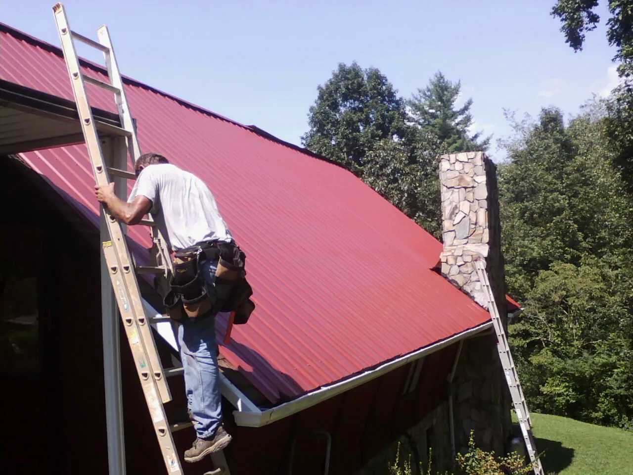 Roofer on a ladder beside a red metal roof and stone chimney — Soffit & Fascia Repair by Roofing Brothers in Franklinton